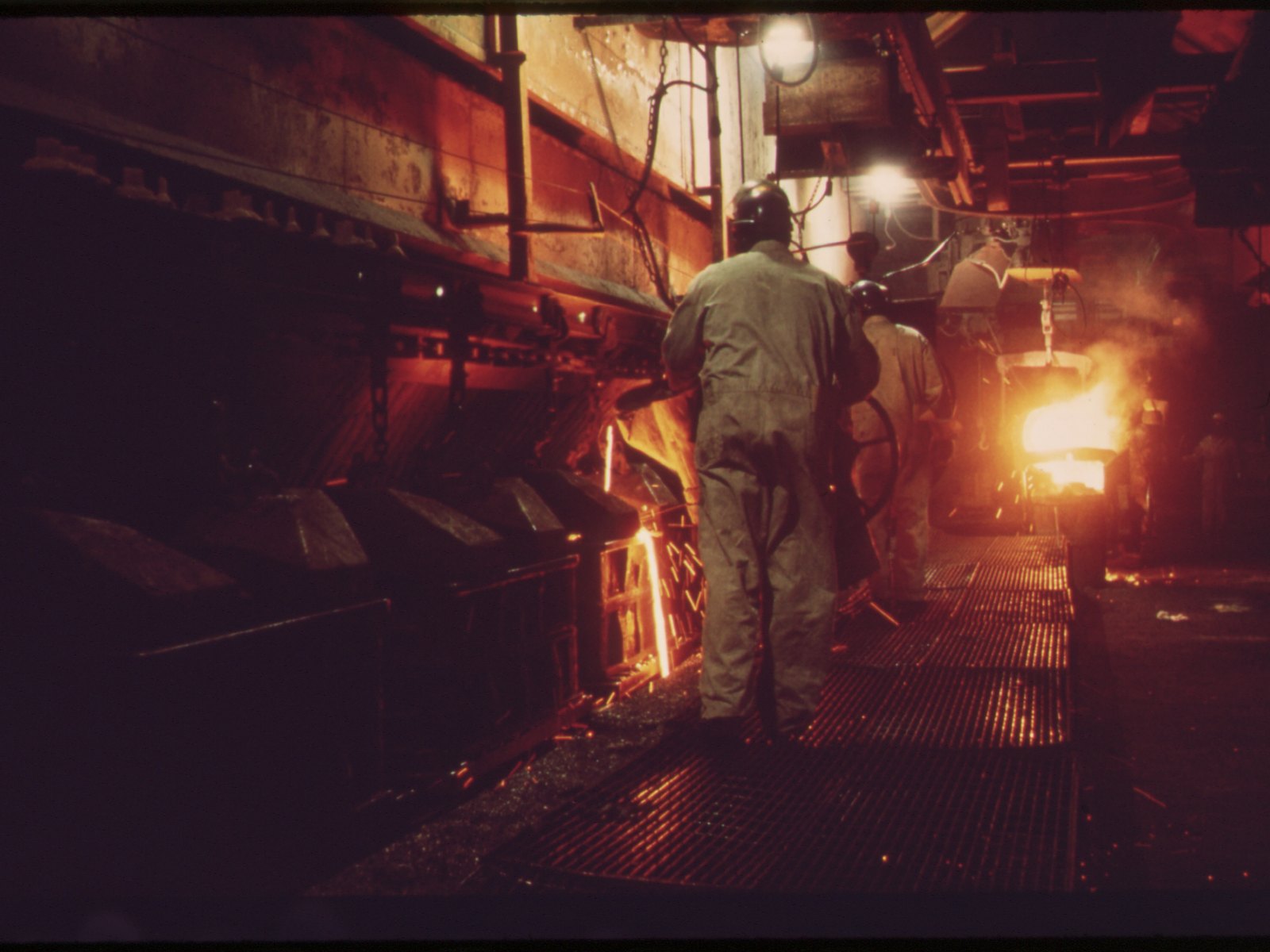 Foundry workers pouring molten metal into a line of sand moulds, sparks at the pour